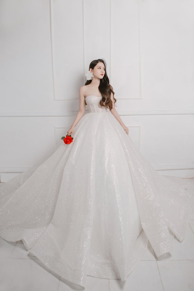 Bride posing with a red rose in luxurious white wedding gown indoors.