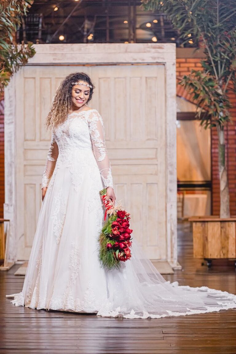 Beautiful bride in lace gown holding vibrant red bouquet indoors with rustic decor.